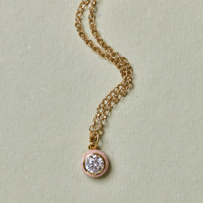 A delicate gold chain necklace with a small round pendant featuring a clear central stone and a soft pink outer bezel, displayed on a textured light green background.