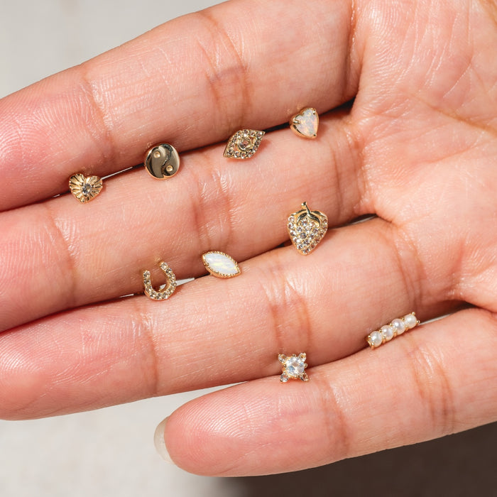 A hand displays eight Solid Gold Opal Marquise Flat Back Studs with various designs—heart, yin-yang, teardrop, horseshoe, pearl row, and gemstones—set against a neutral background.